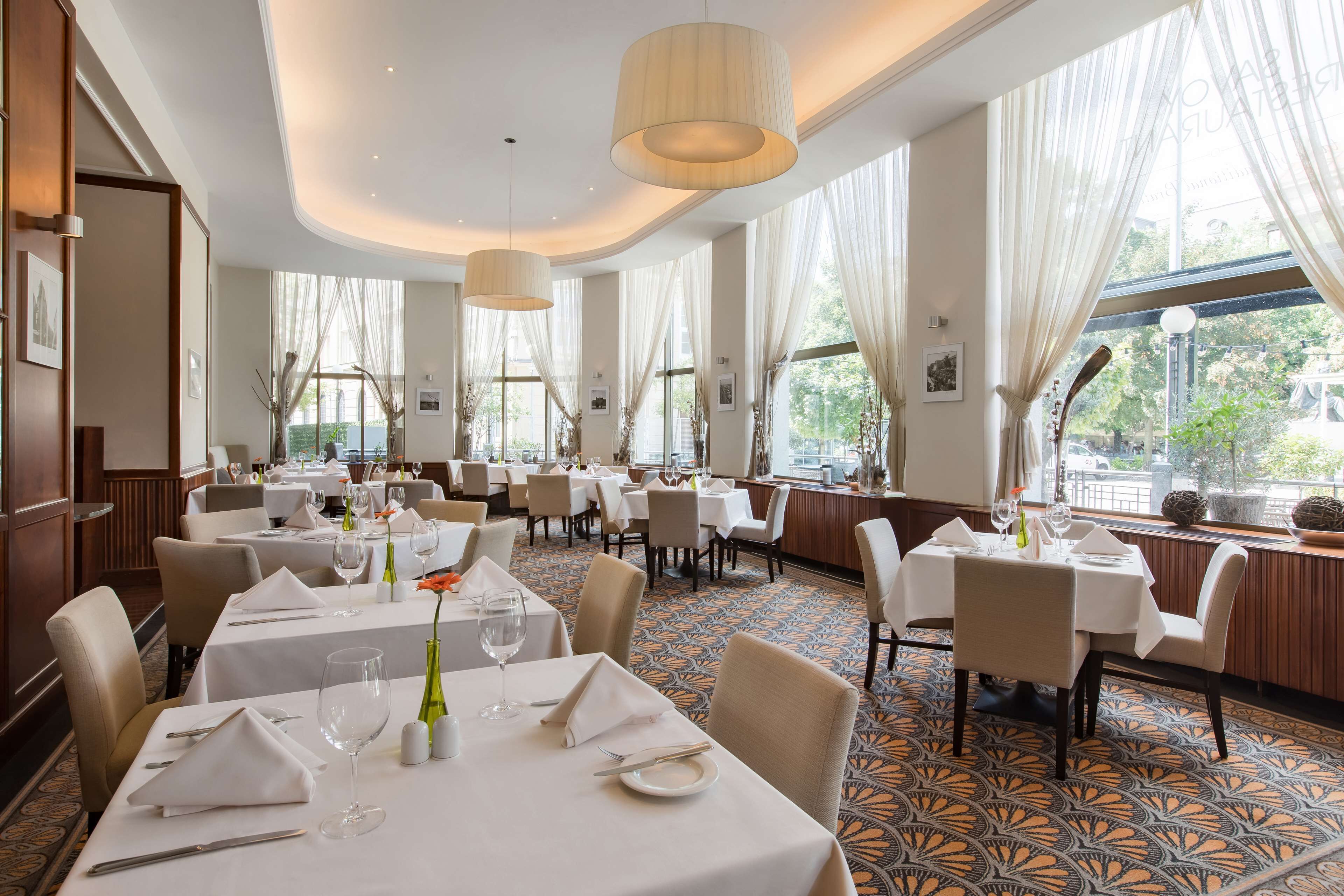 A spacious dining area with multiple tables set for dining, large windows with sheer curtains, and a patterned carpet.