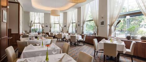 A spacious dining area with multiple tables set for dining, large windows with sheer curtains, and a patterned carpet.