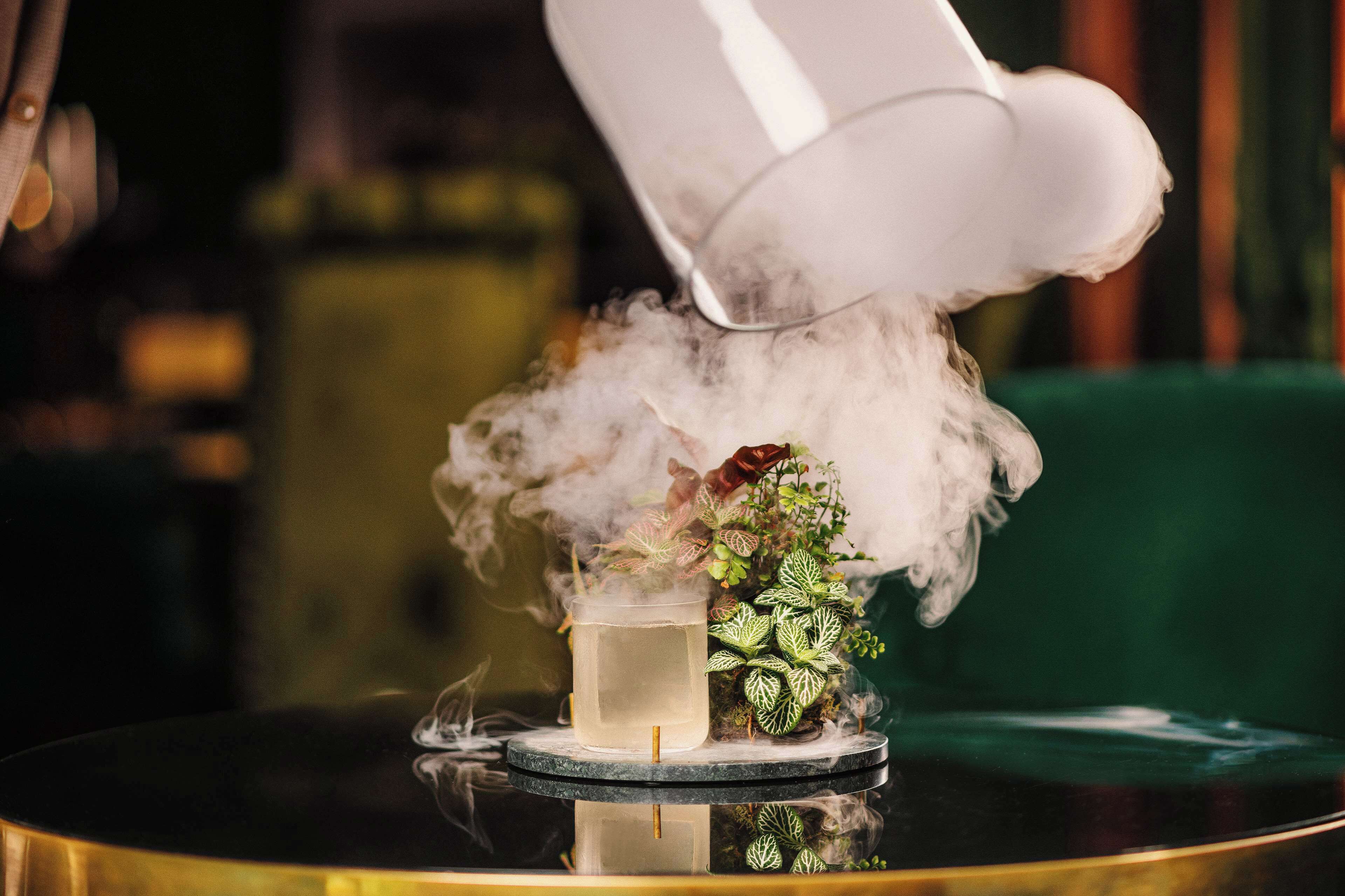 A cocktail with a glass on a round table, surrounded by a decorative plant and a white object emitting steam.