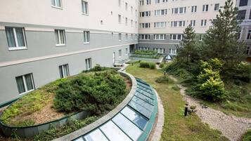 Un patio con jardín en la azotea verde, un pasillo curvo de vidrio y un edificio con múltiples ventanas.