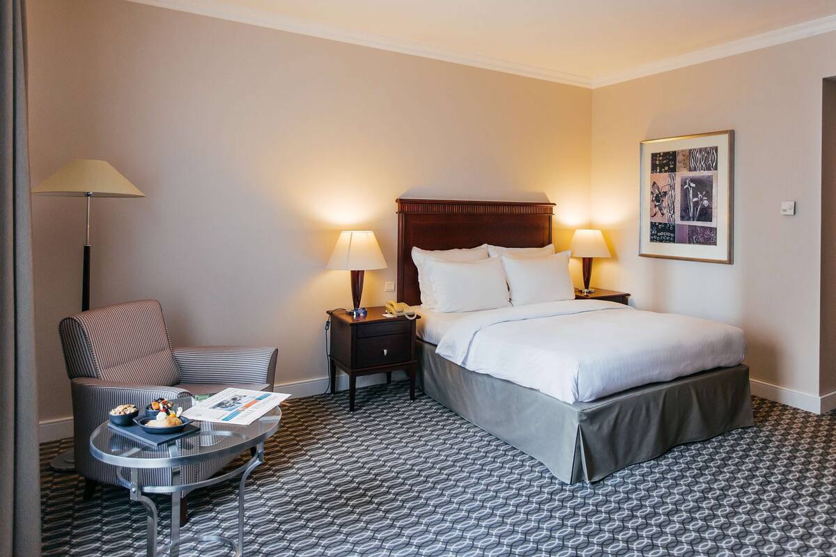A hotel room with a bed, bedside tables, a chair, a small table with a lamp, and a patterned carpet.