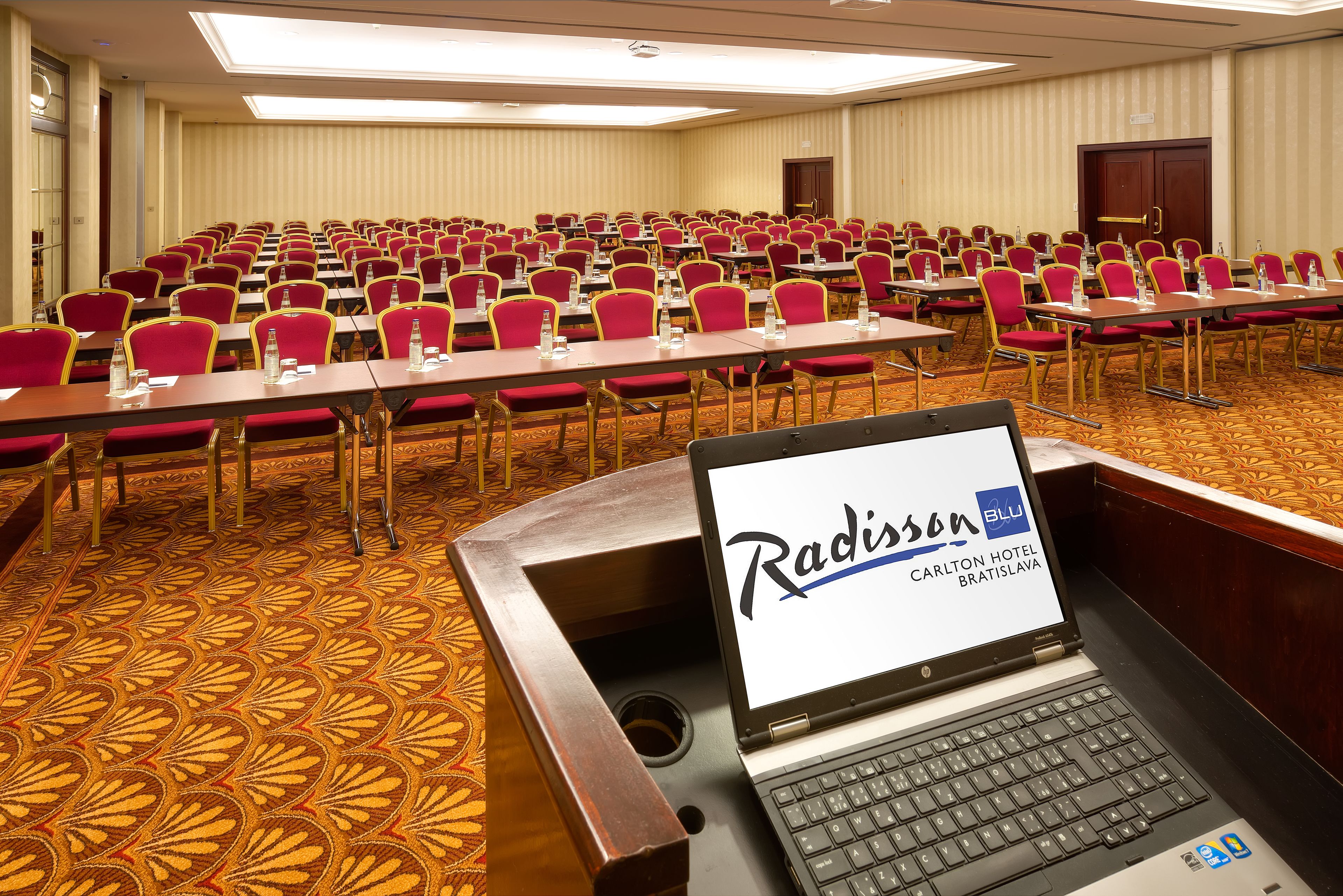 A conference room with rows of red chairs and tables, a laptop displaying 'Radisson Blu Carlton Hotel Bratislava', and a carpeted floor.