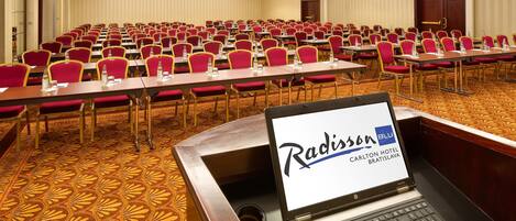 Un salón de conferencias con filas de sillas rojas y mesas, una laptop mostrando 'Radisson Blu Carlton Hotel Bratislava', y piso alfombrado.