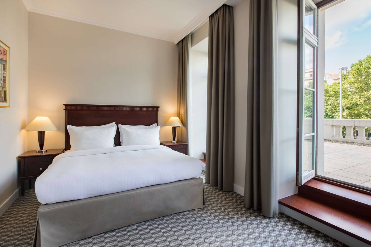 A hotel room with a large bed, two bedside tables with lamps, a window with curtains, and a patterned floor.