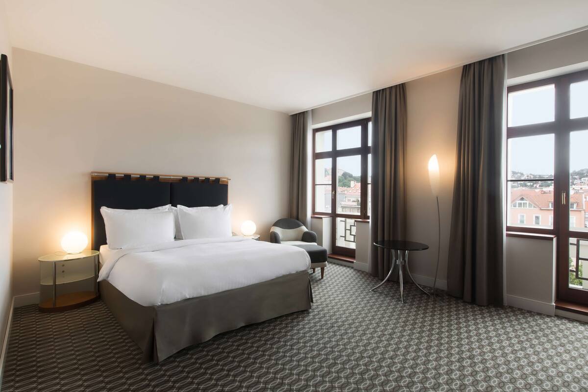 A hotel room with a large bed, a chair, a small table, and a view of the city through large windows.