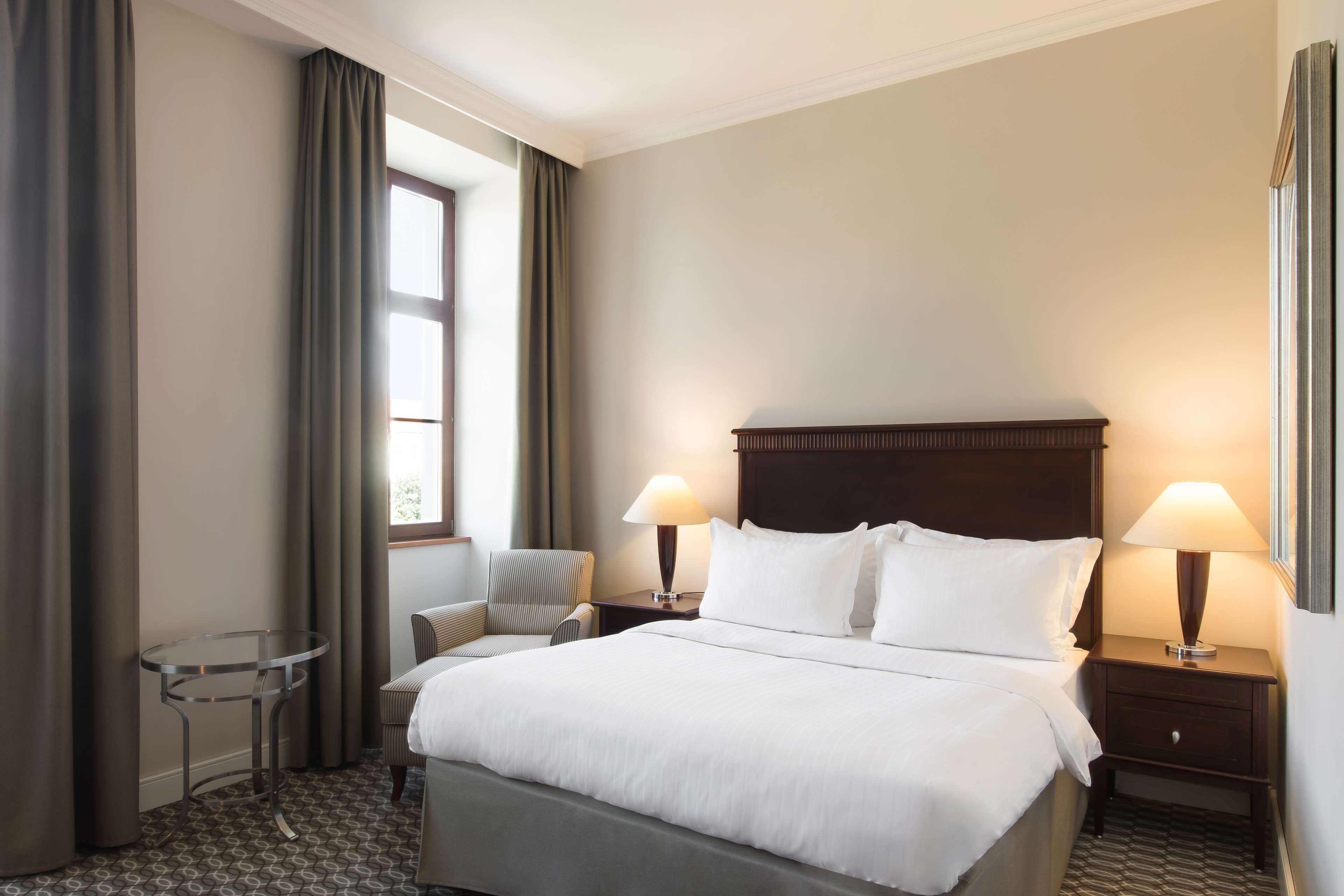 A hotel room with a large bed, two bedside lamps, a chair, a small table, and a window with curtains.