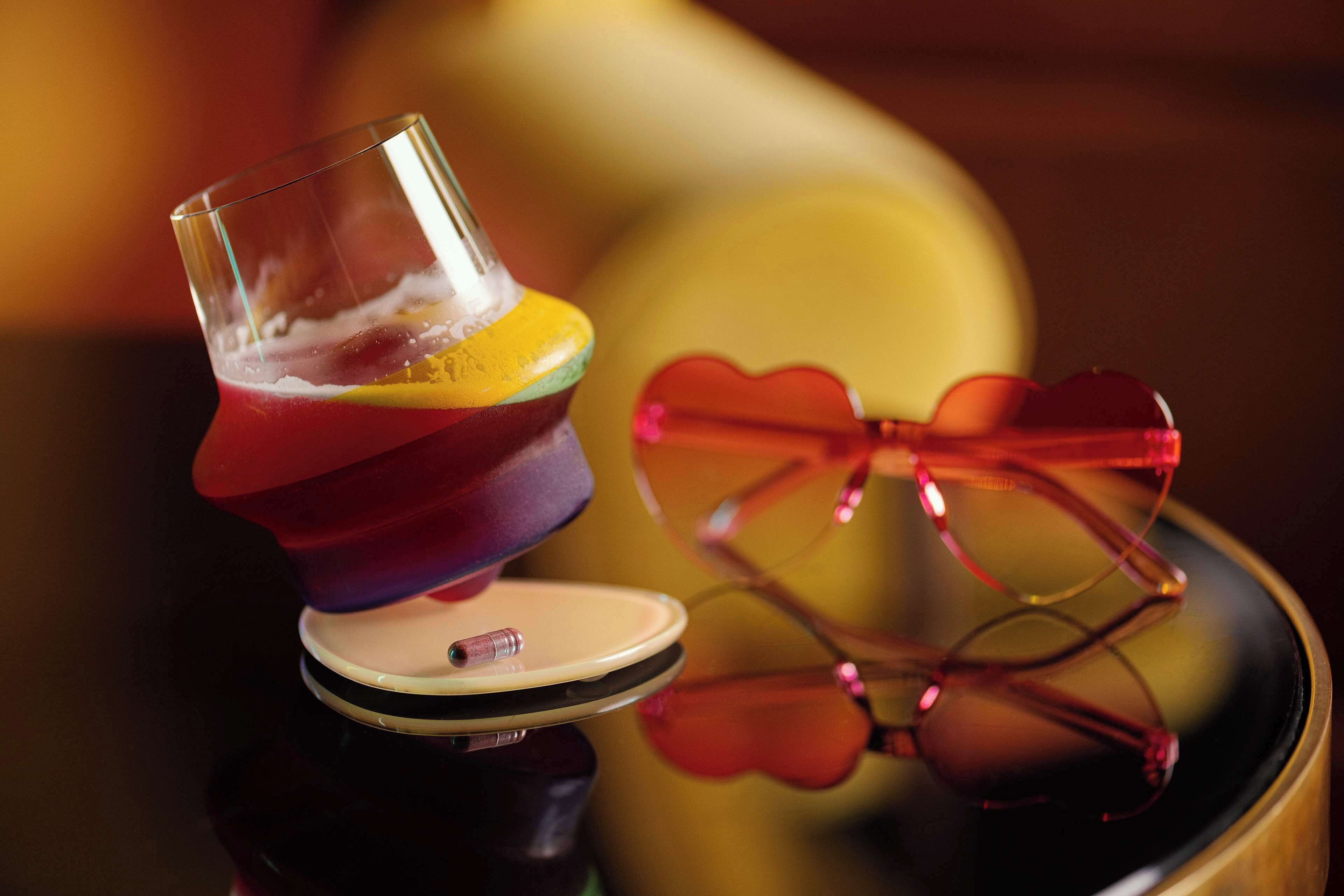 A cocktail glass with a layered drink, a pair of heart-shaped sunglasses, and a blurred background.