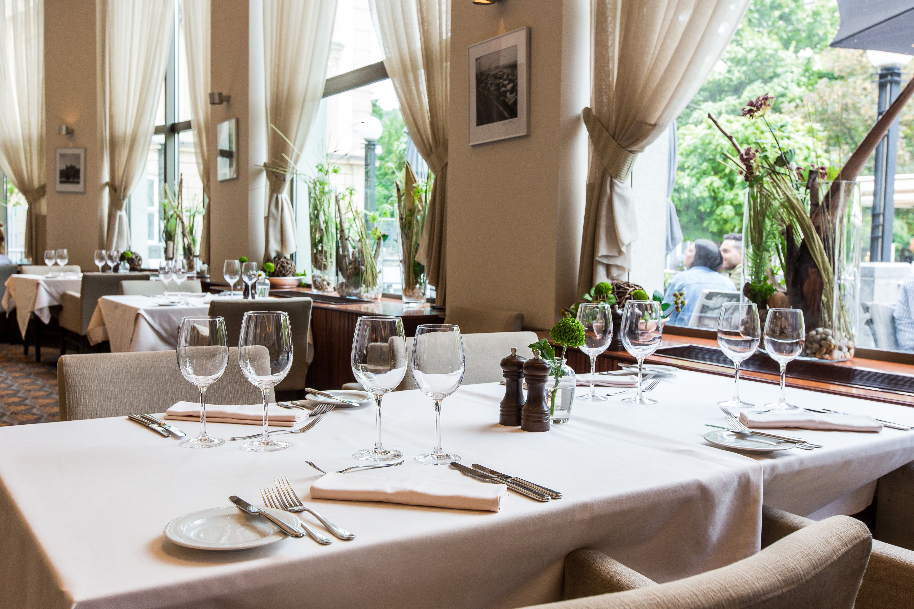 Un comedor con mesas dispuestas para una comida, con manteles blancos, cristalería y cubiertos ordenados.