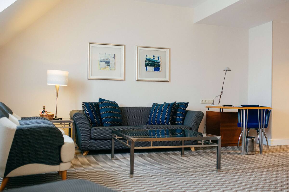 A modern living room with a sofa, armchair, coffee table, and a desk with a chair. There are two framed pictures on the wall.
