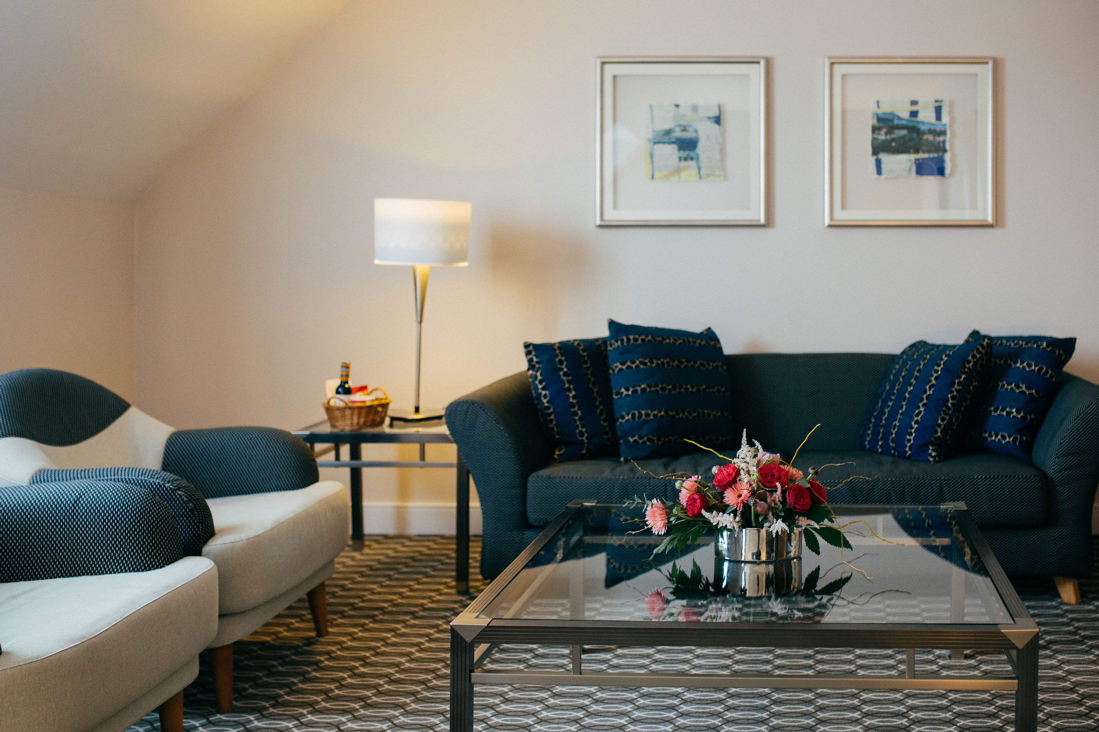 Een woonkamer met een bank, fauteuils, een bijzettafeltje met bloemen, een lamp en twee ingelijste foto's aan de muur.