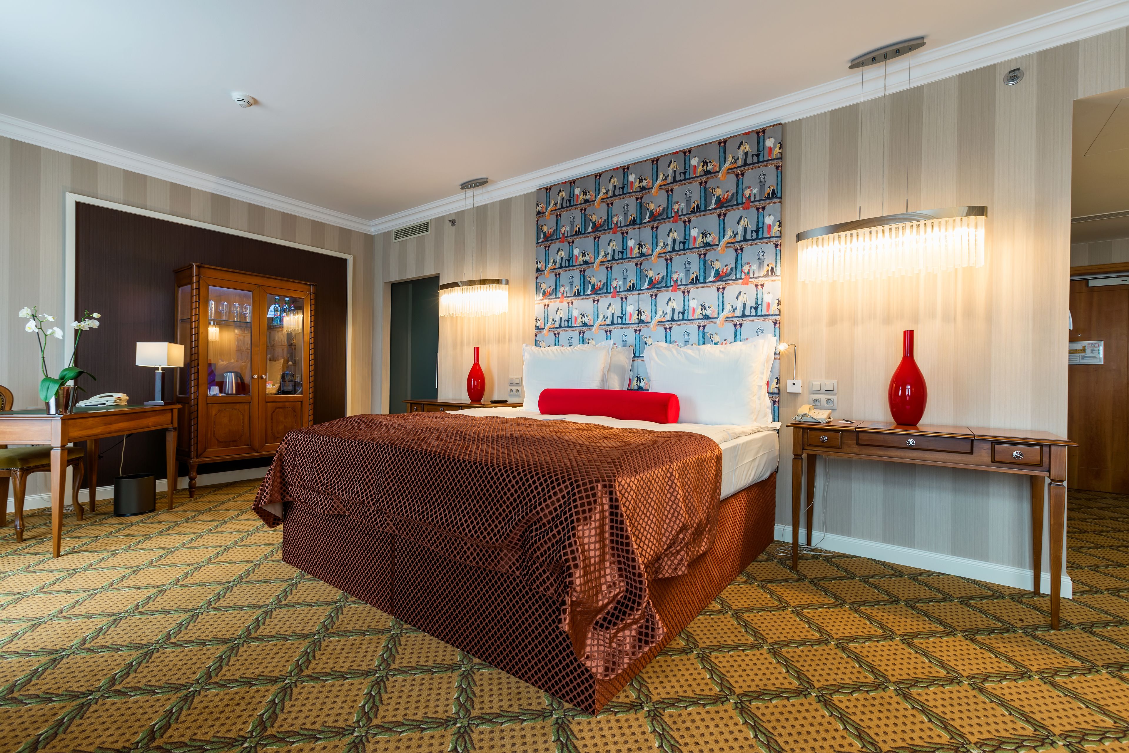 A hotel room with a large bed, a desk, a chair, a wardrobe, and a patterned carpet.