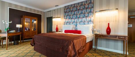 A hotel room with a large bed, a desk, a chair, a wardrobe, and a patterned carpet.