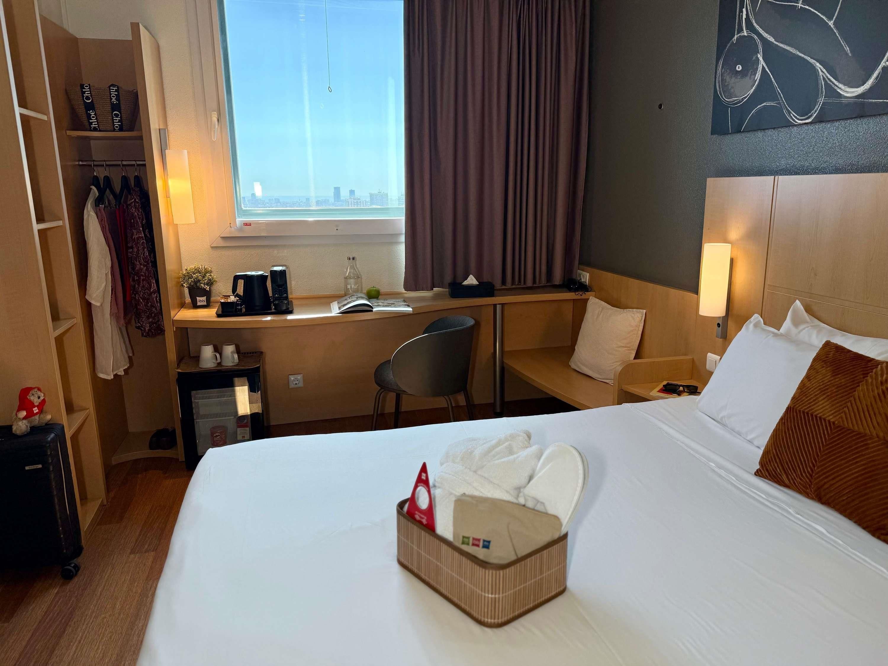 A hotel room with a bed, a desk, a chair, a wardrobe, and a view of the city.