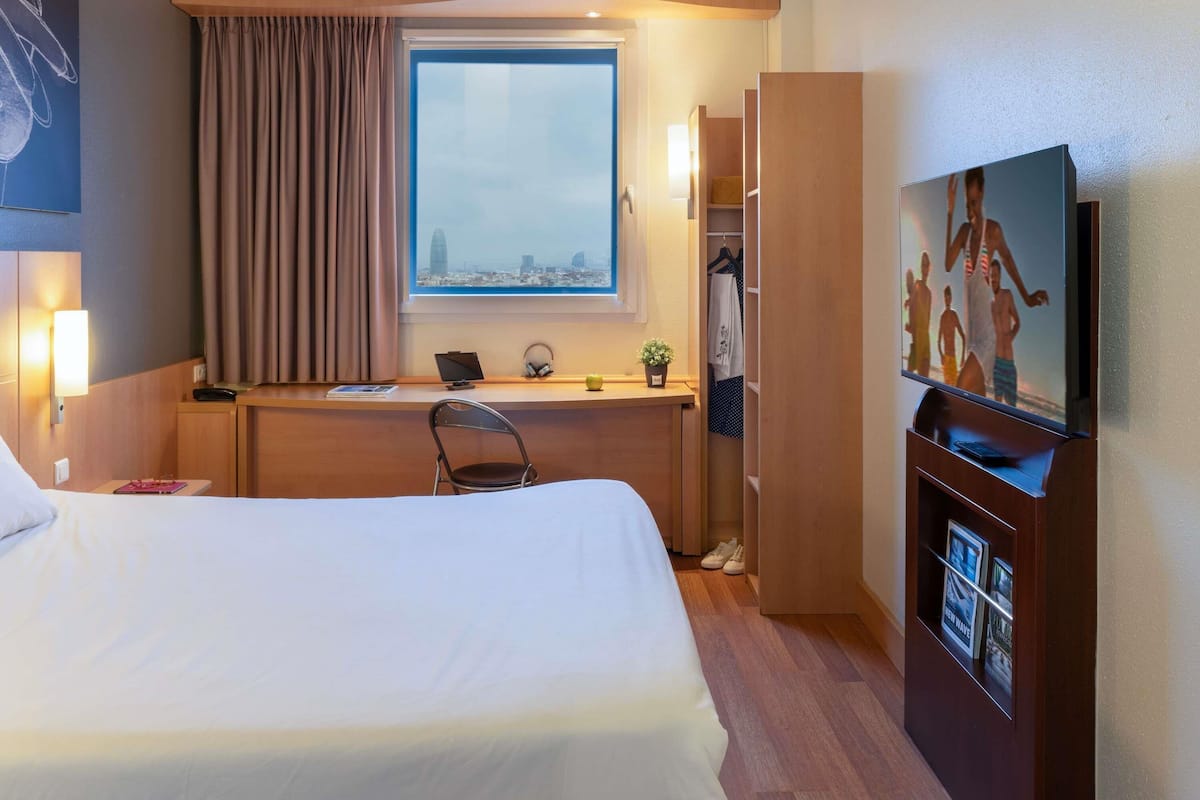 Habitación de hotel con cama, escritorio con laptop, televisión y ventana con vista a la ciudad.