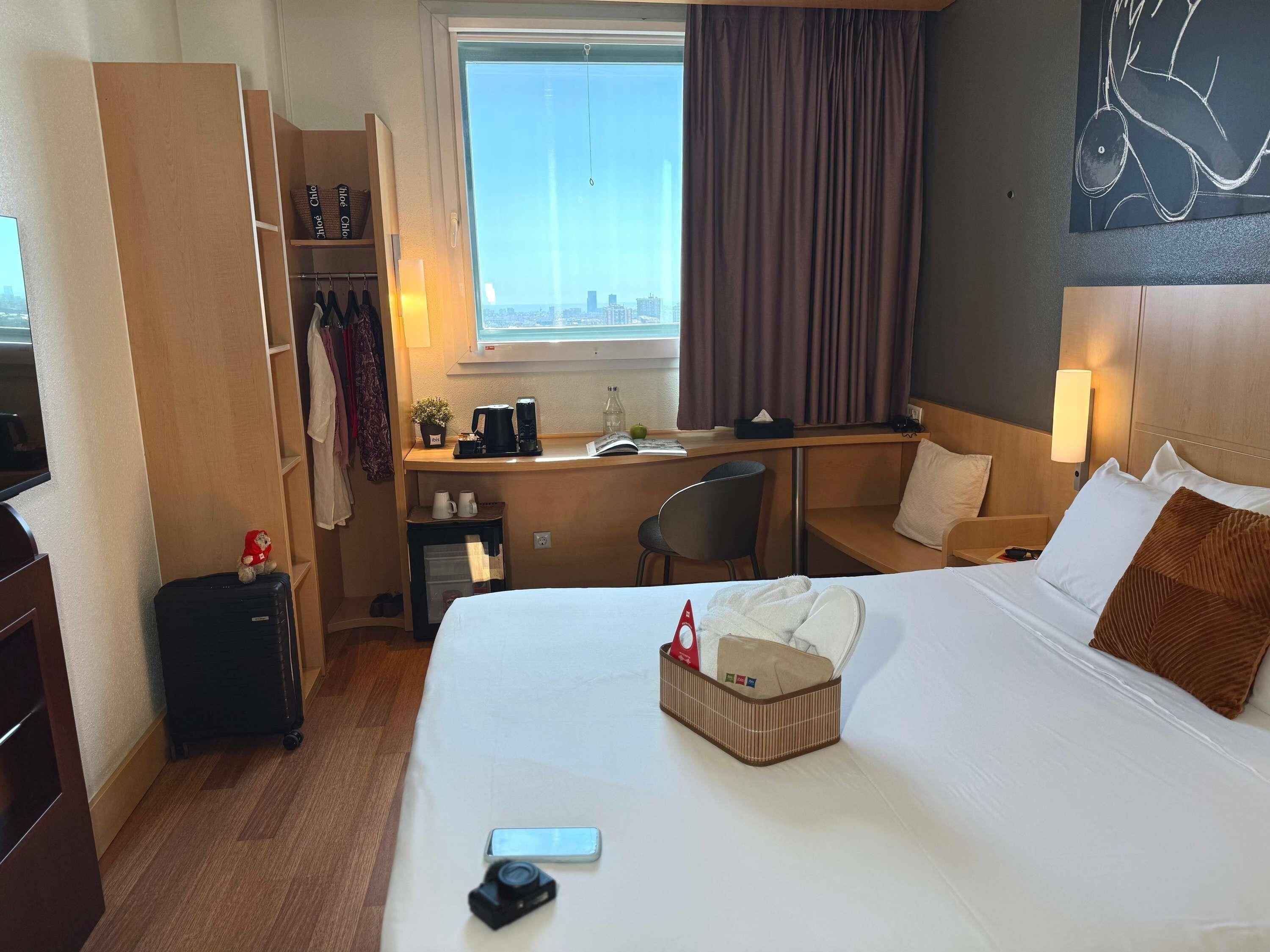 A hotel room with a bed, desk, chair, wardrobe, and a view of the city.