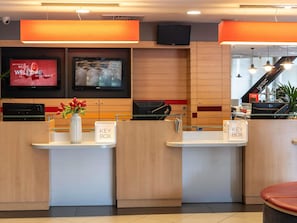 Zona de recepción de un hotel con paneles de madera, un televisor Sony mostrando un cartel de película, un buzón de llaves y un mostrador de recepción.