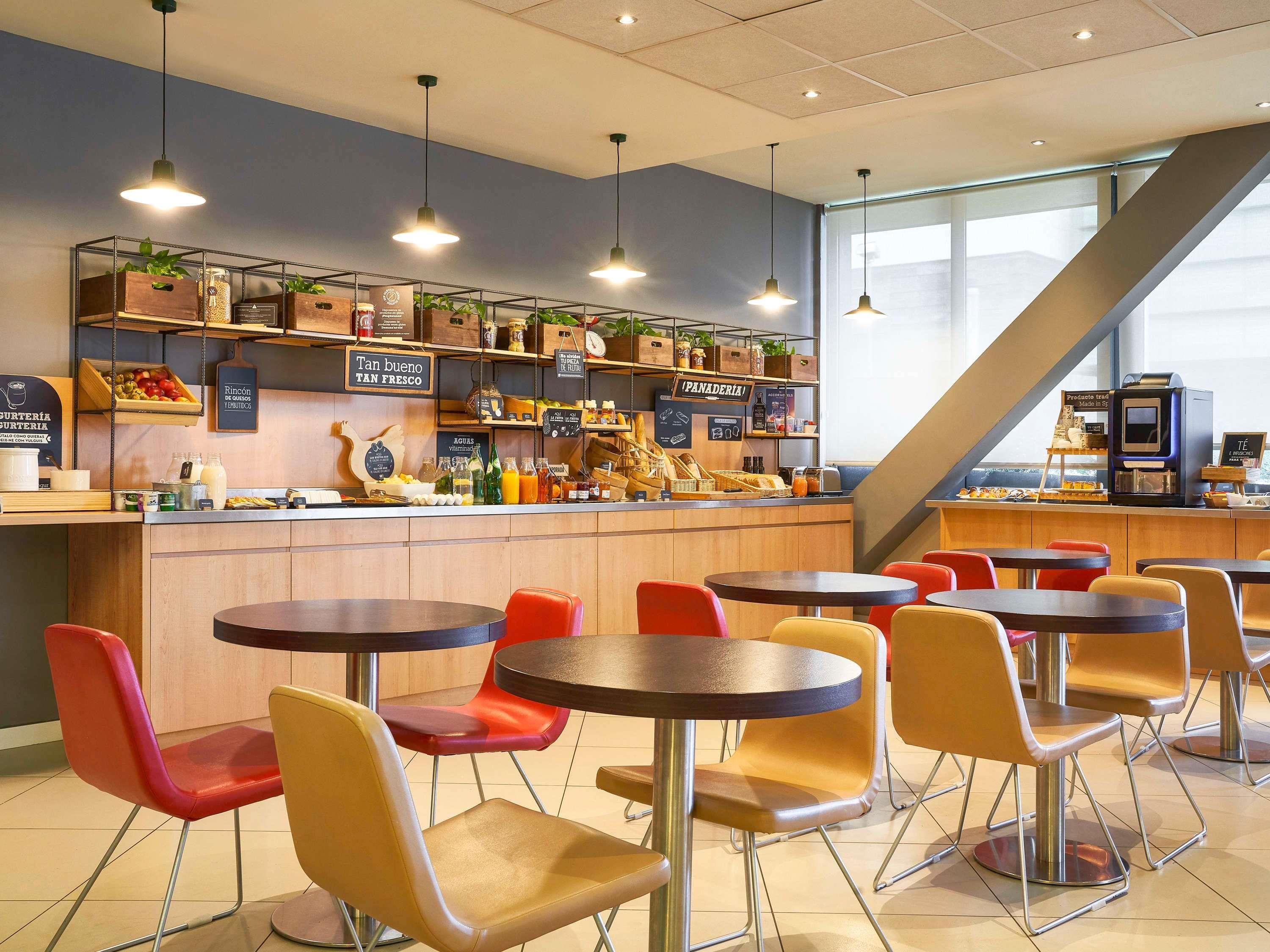 Un café avec plusieurs tables et chaises, un comptoir où sont exposés divers produits alimentaires, et un escalier en arrière-plan.