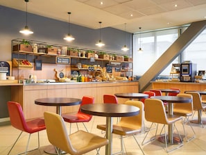 Una cafetería con varias mesas y sillas, una barra con diferentes productos alimenticios y una escalera al fondo.