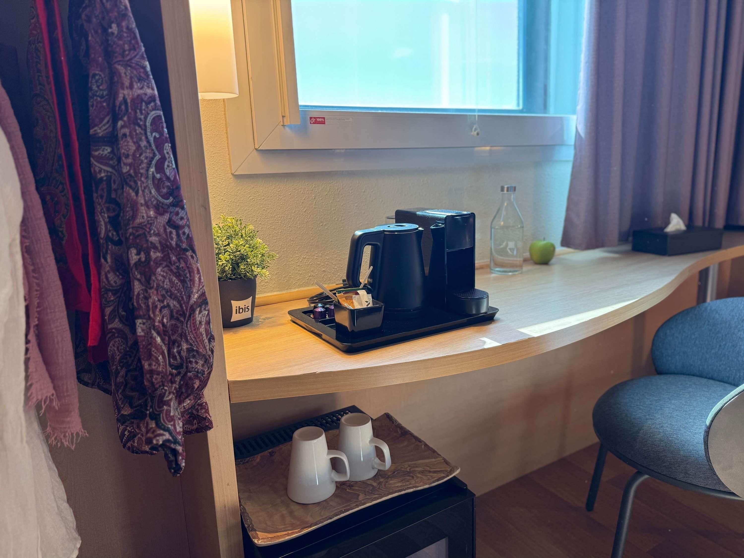 A hotel room with a coffee maker, a small table with mugs, a potted plant, and a window with curtains.