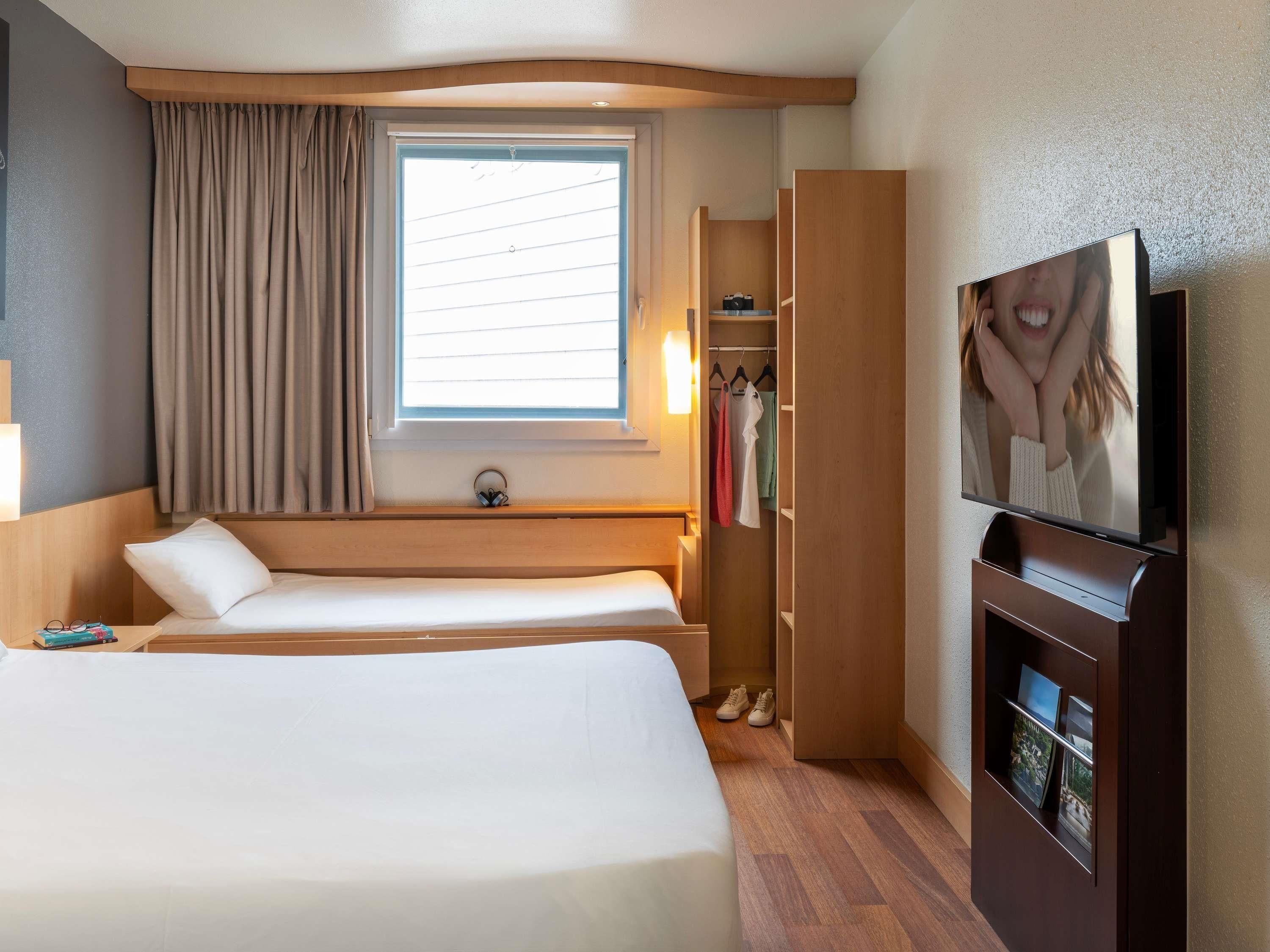 A hotel room with two beds, a window with curtains, a TV, a wardrobe, and a shoe rack.