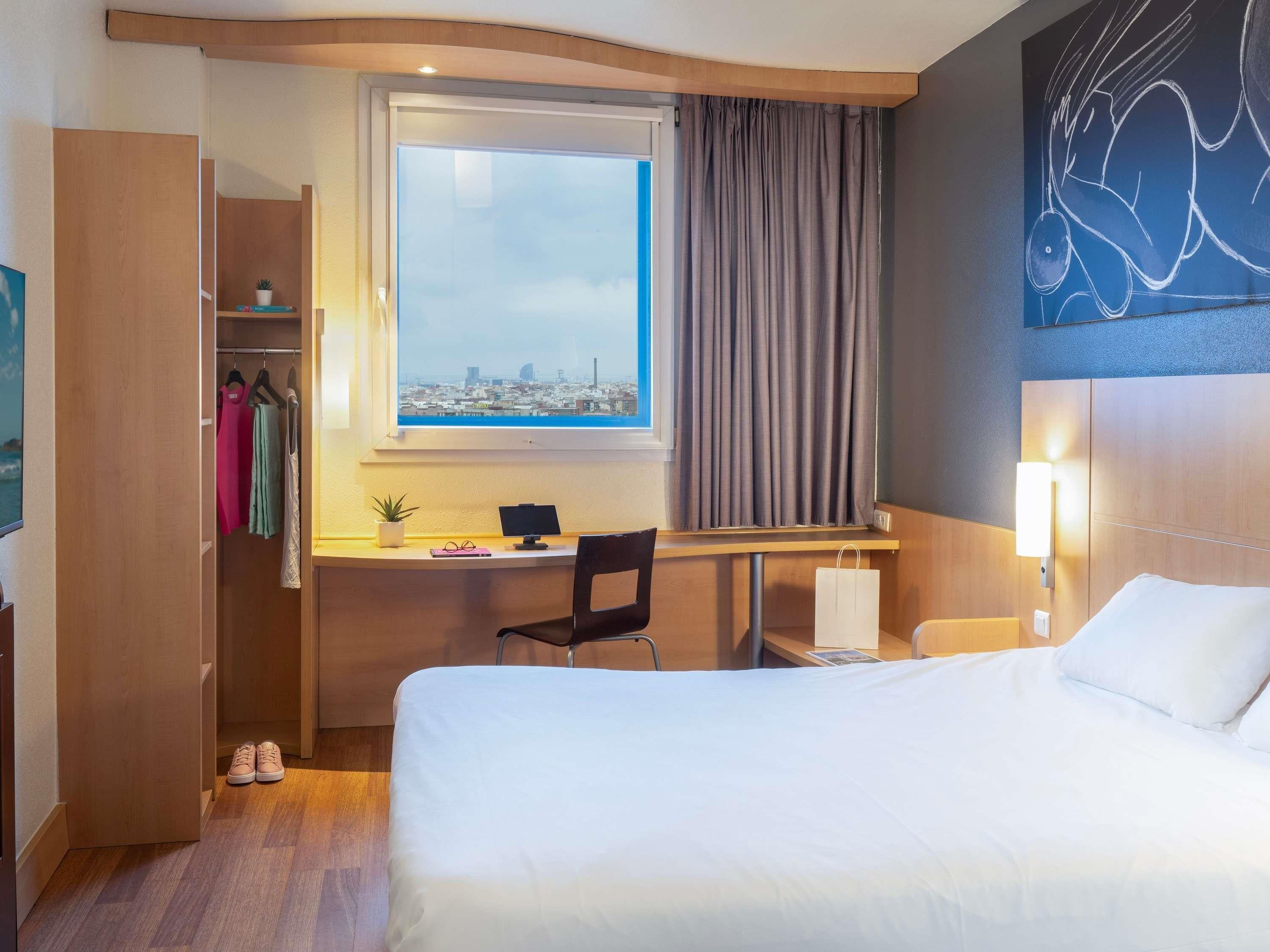 Hotelkamer met een bed, bureau met een computer, stoel, kast en uitzicht op de stad door een raam.