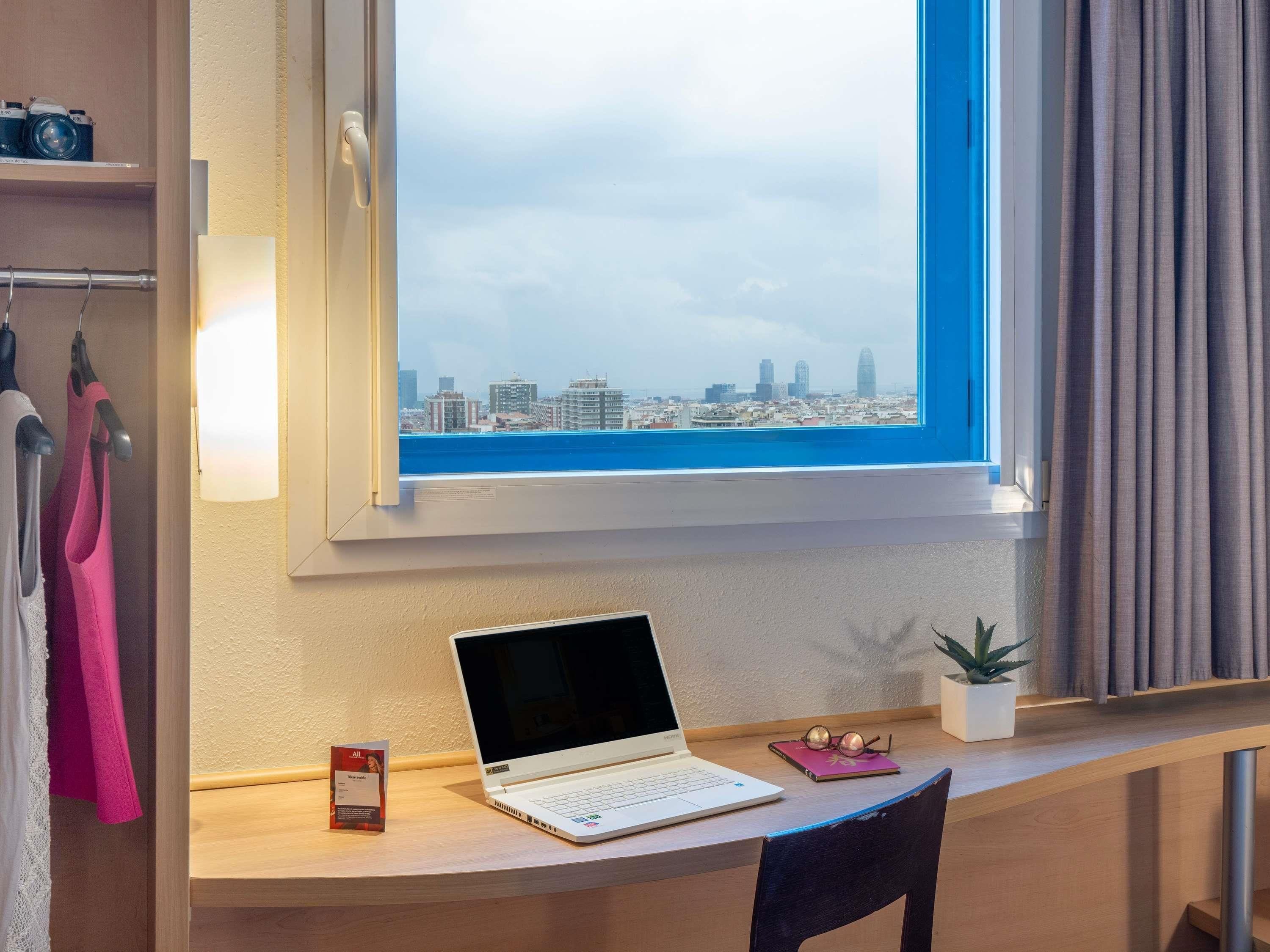A room with a desk, a laptop, a small plant, and a window with a city view.