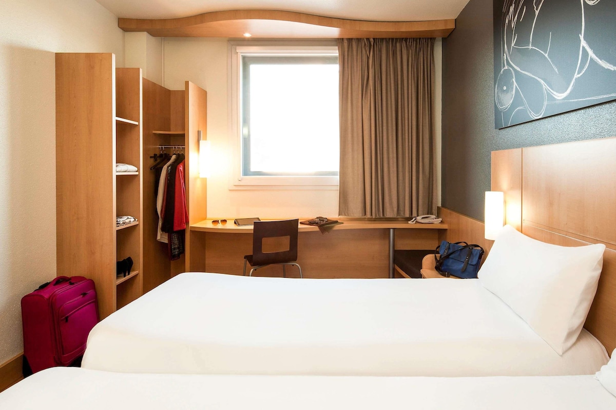 A hotel room with a bed, desk, chair, wardrobe, and a window with curtains.
