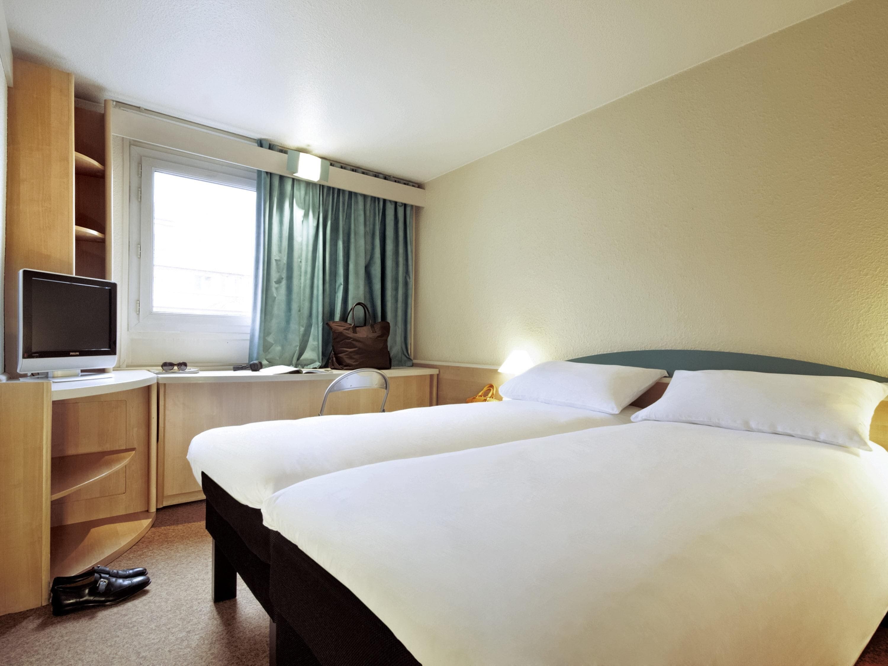 Una habitación de hotel con una cama, un escritorio con monitor de computadora, una silla y un ventanal con cortinas.