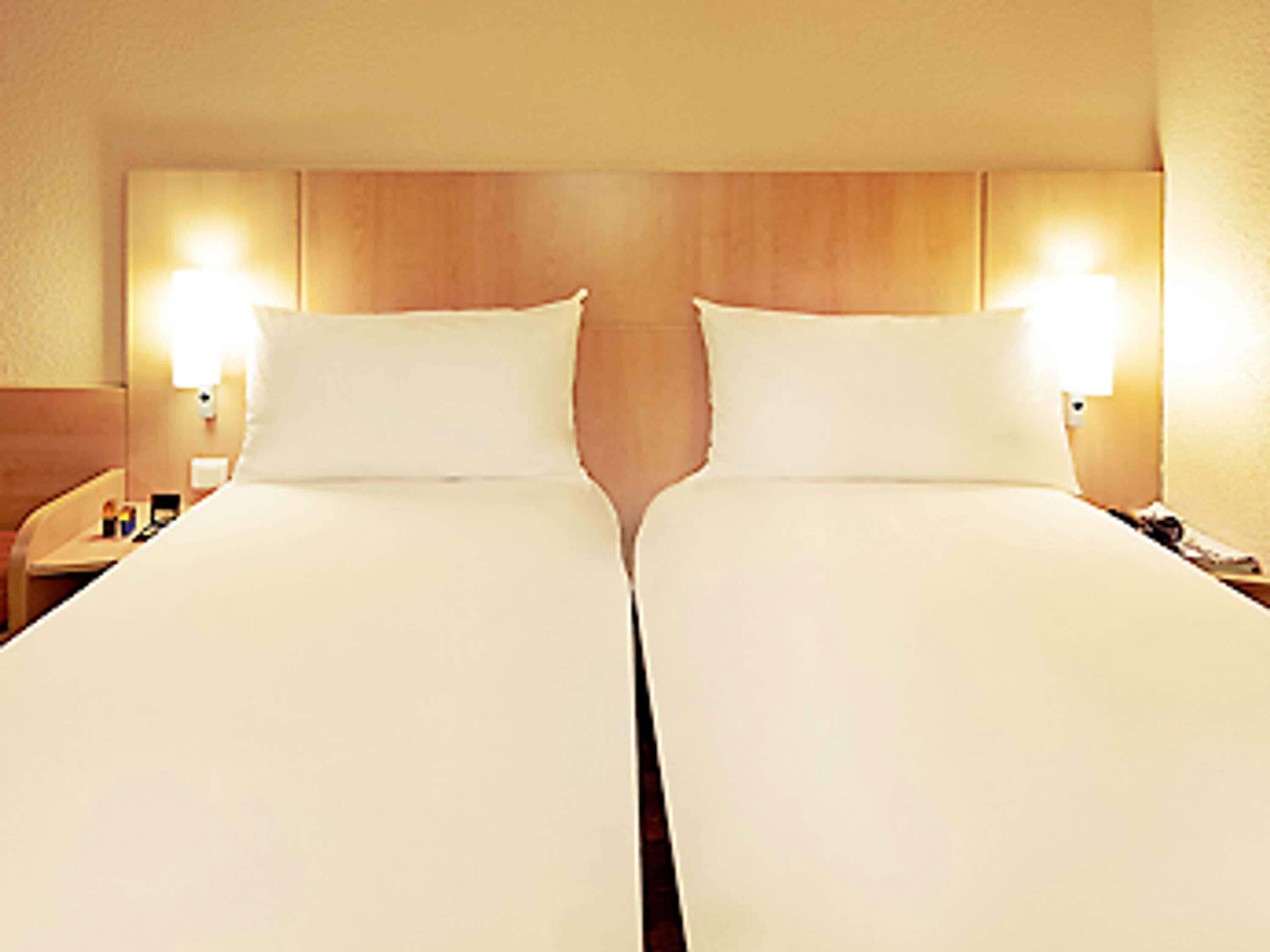 A hotel room with two single beds, wooden headboard, bedside lamps, and a nightstand with a lamp.