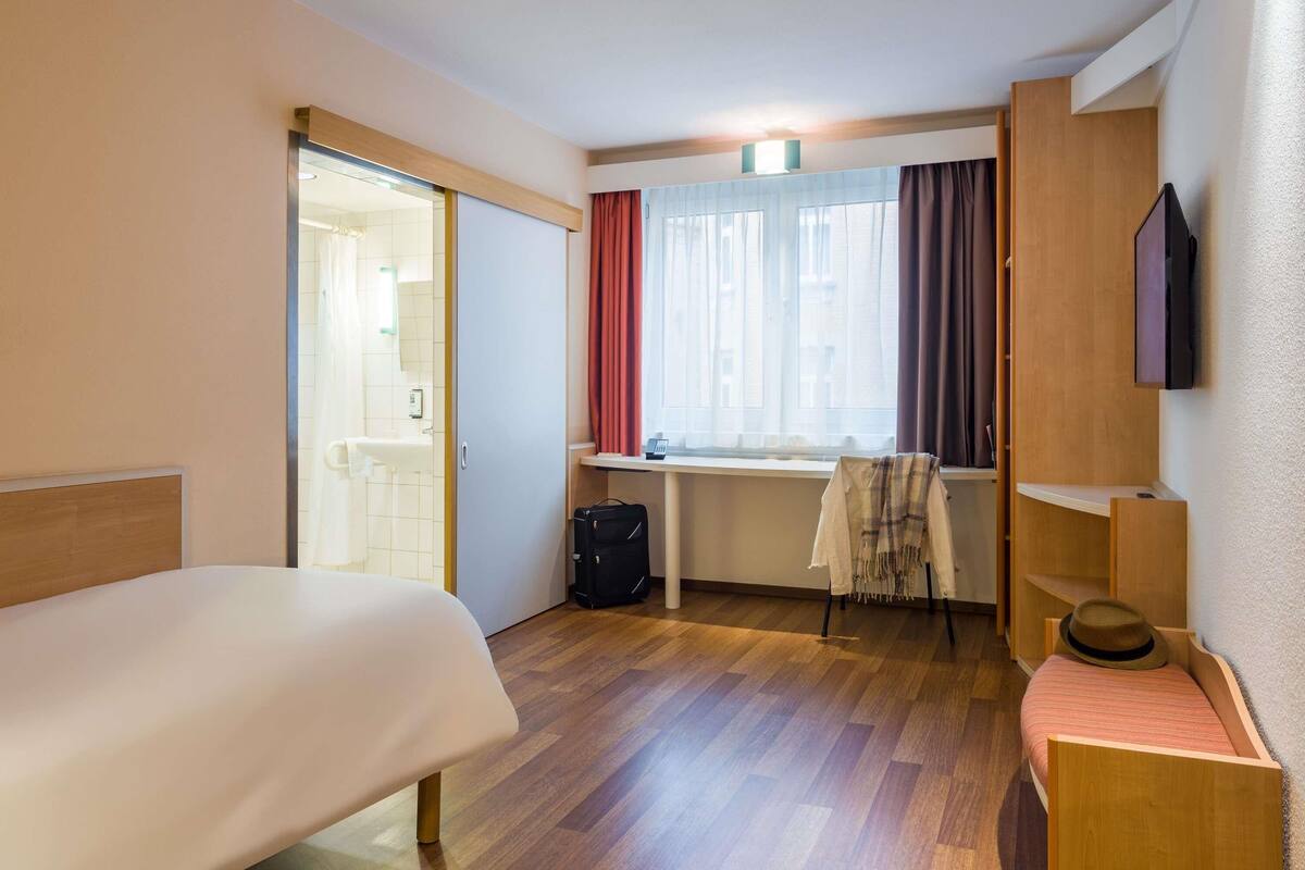 A hotel room with a bed, a desk, a chair, a suitcase, and a TV.