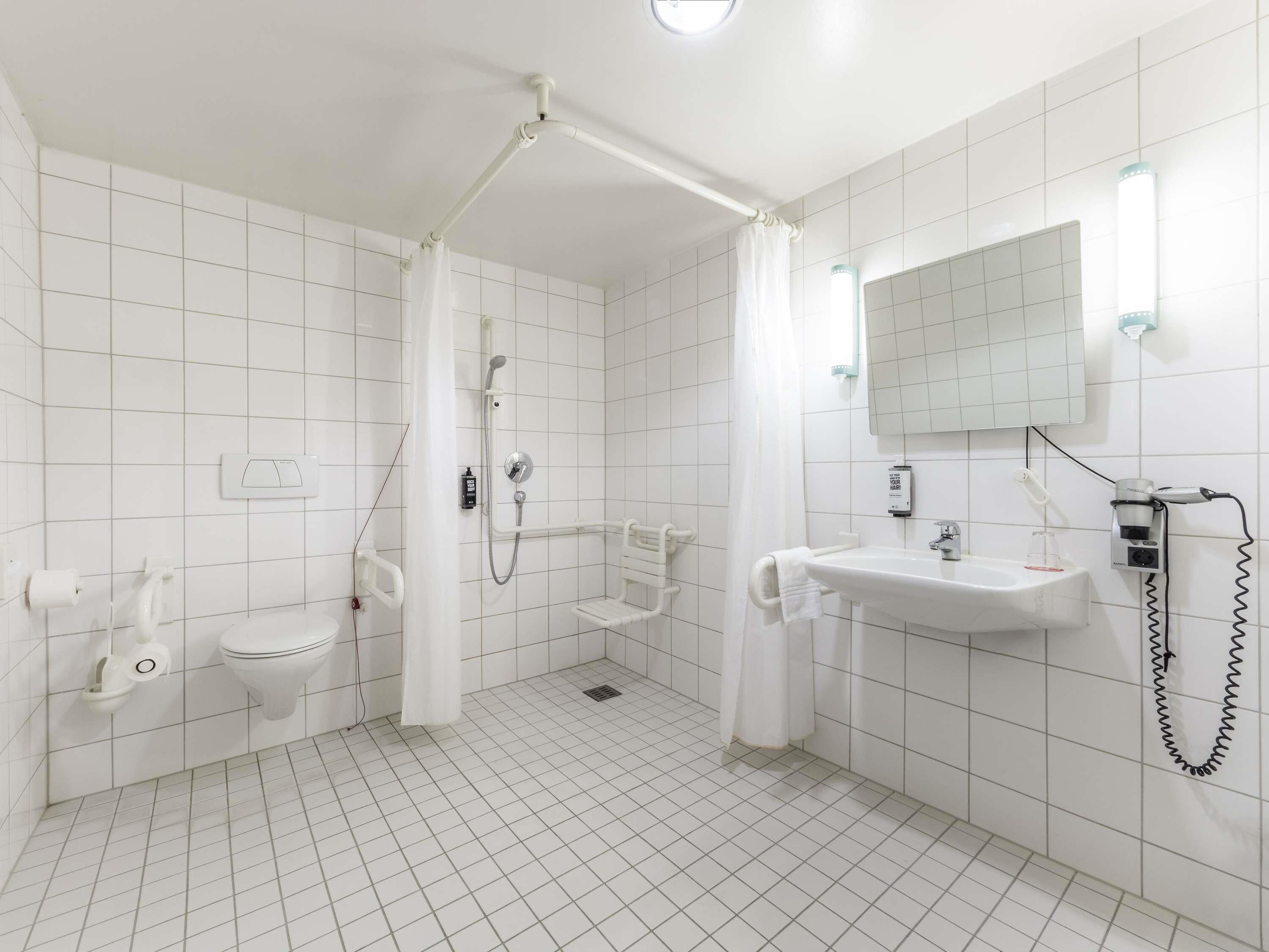 A bathroom with white tiles, a shower, a toilet, a sink, and a mirror.