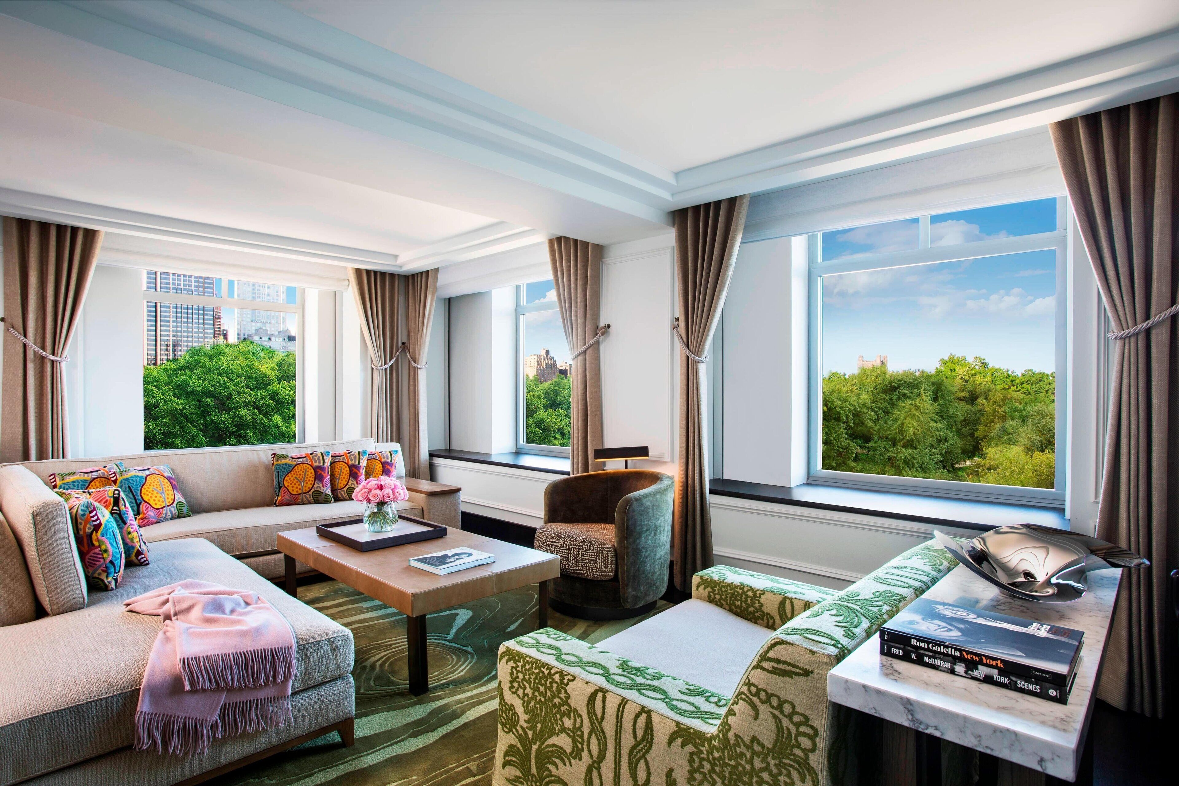 A modern hotel room with a large window, a sofa, a chair, a coffee table, and a small table with books.