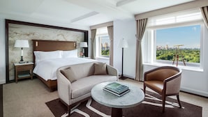 A hotel room with a large bed, two bedside tables with lamps, a sofa, a small round table, and a view of a cityscape through large windows. - The Ritz-Carlton New York, Central Park (New York)