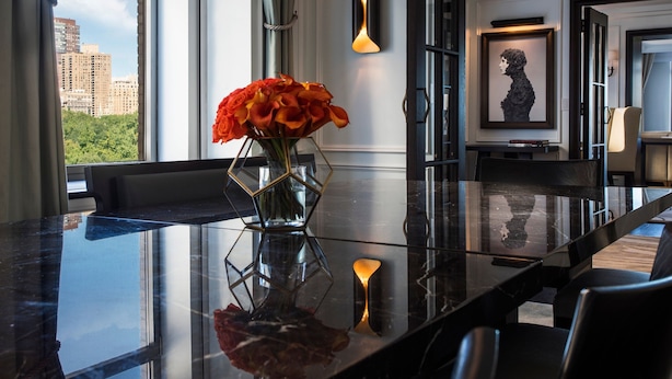 A modern dining area with a glass table, a vase of flowers, and a city view through large windows. - The Ritz-Carlton New York, Central Park (New York)