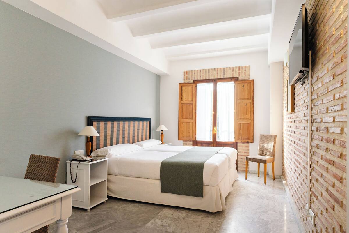 A hotel room with a bed, a desk, a chair, a TV mounted on a brick wall, and a window with wooden shutters.