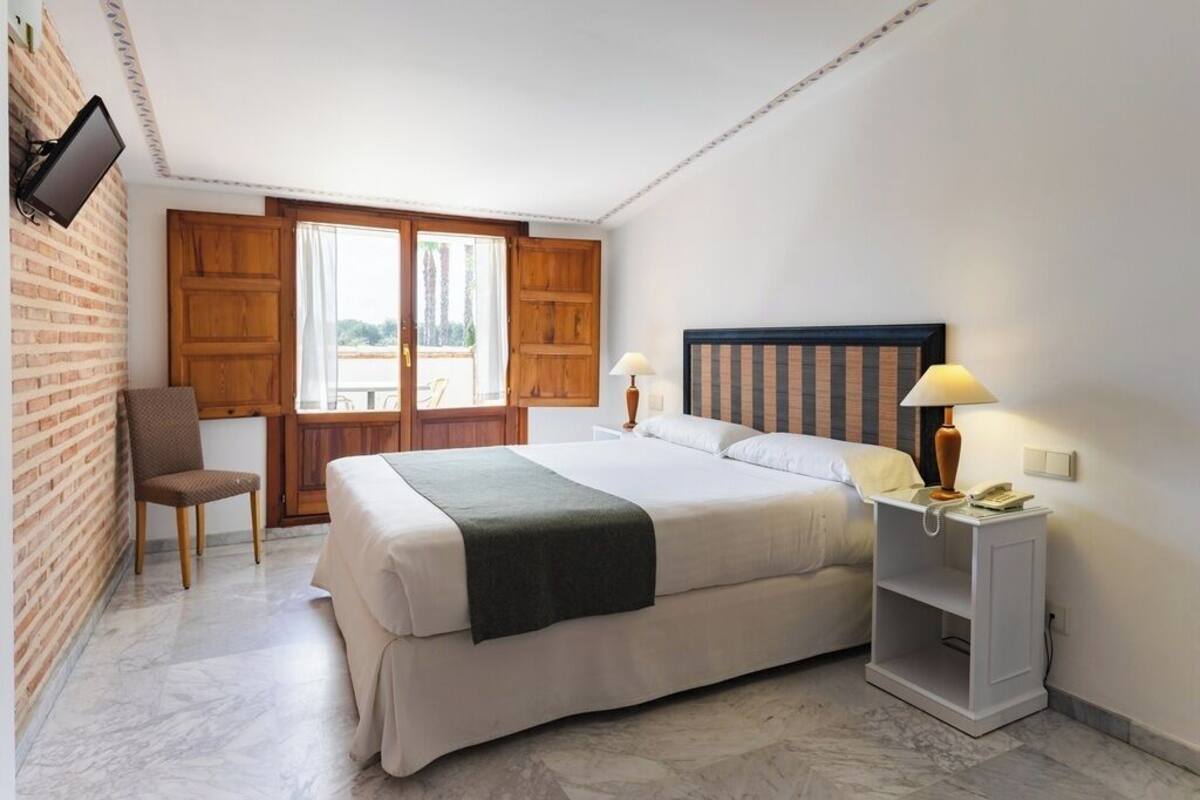 A bedroom with a large bed, a chair, a nightstand with a lamp, and a TV mounted on the wall.