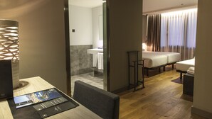 A hotel room with a wooden floor, a desk with a chair, a bed with two pillows, a bedside table, and a window with curtains.