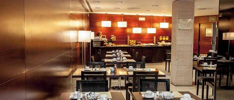 A hotel dining area with multiple tables set for breakfast, a buffet counter in the background, and a modern interior design with wooden walls and ceiling lights.