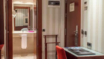 A hotel room with a wooden floor, a bed, a desk with a chair, a television, and a bathroom visible through an open door.