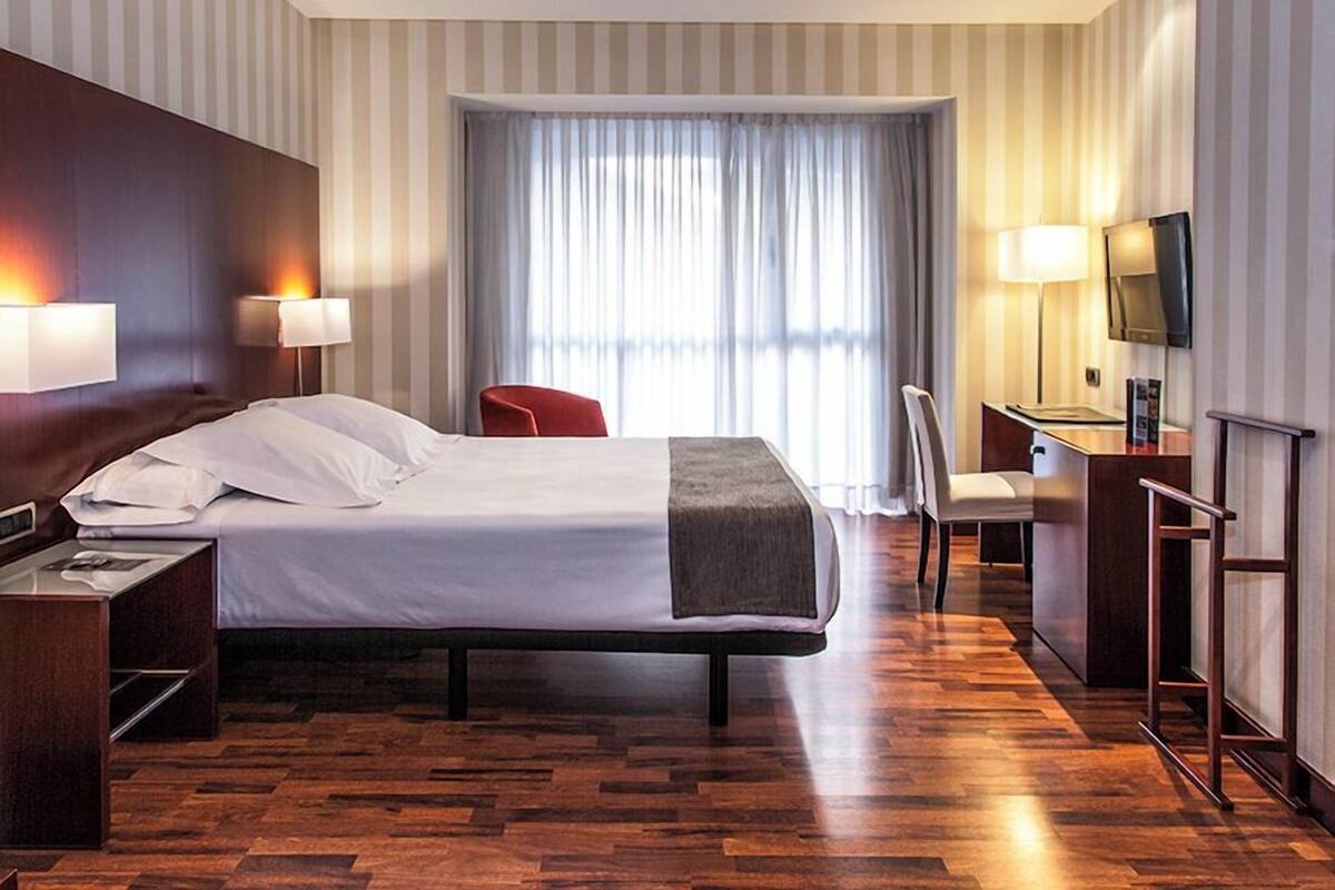 Ein Hotelzimmer mit Bett, Nachttischen, einem Schreibtisch mit Stuhl, einem Fernseher und einem Fenster mit Vorhängen.