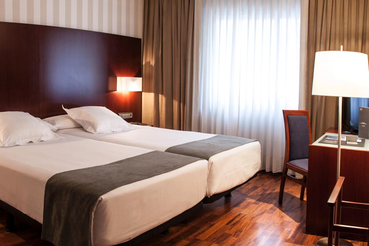 Quarto de hotel com duas camas, uma escrivaninha, uma cadeira e um abajur.