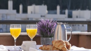 A breakfast table with croissants, a coffee pot, and two glasses of orange juice, set against a backdrop of trees and buildings.