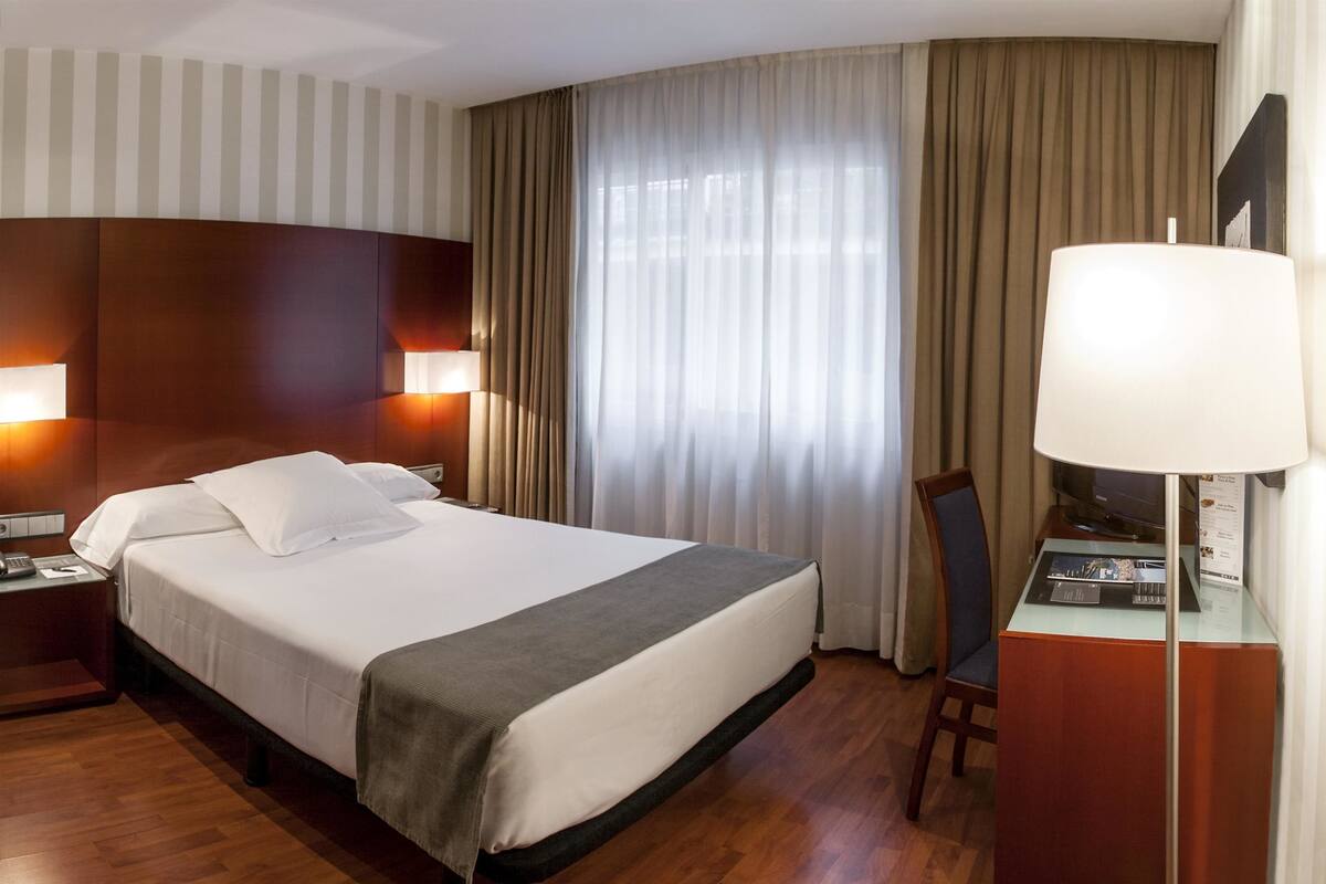 Ein Hotelzimmer mit einem großen Bett, einem Schreibtisch mit Lampe, einem Sessel und einem Fenster mit Vorhängen.