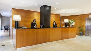 A reception desk with two staff members, a wooden panel wall, a potted plant, and an elevator.
