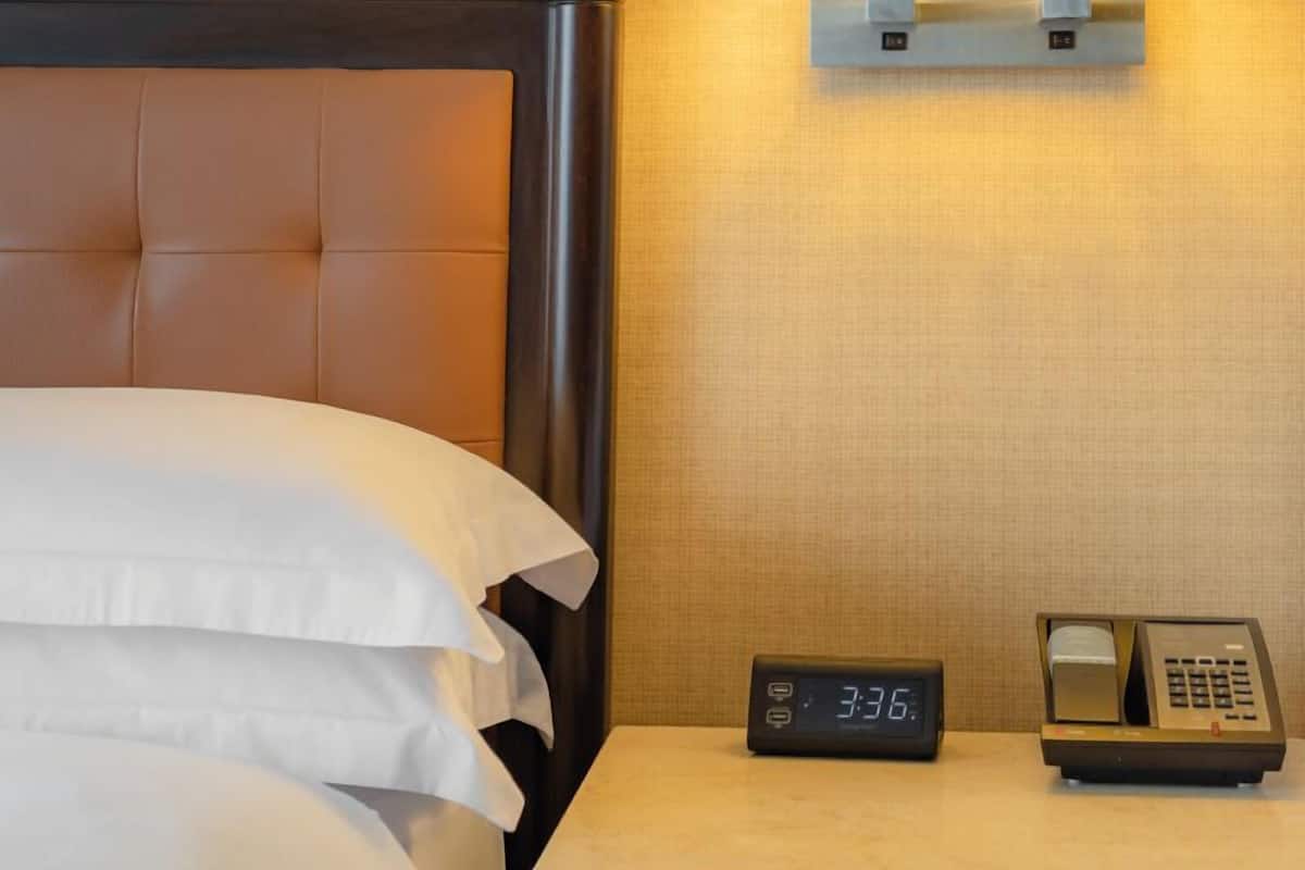 A hotel room with a bed, a nightstand with a lamp, a digital clock, and a telephone.