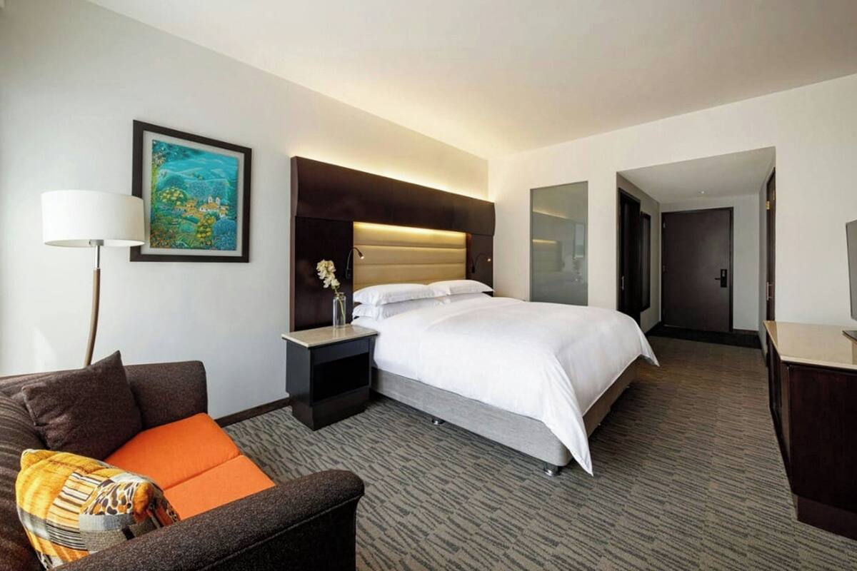 A hotel room with a bed, a nightstand, a lamp, a sofa, and a painting on the wall.