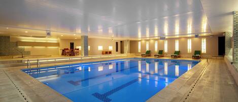 Una piscina interior con carriles delimitados, sillas de descanso y una zona de recepción con mostrador y sillas.