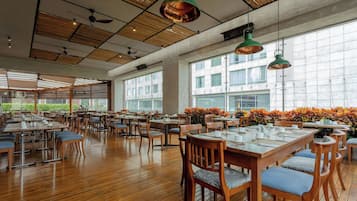 A spacious dining area with wooden tables and chairs, large windows, and hanging green pendant lights.