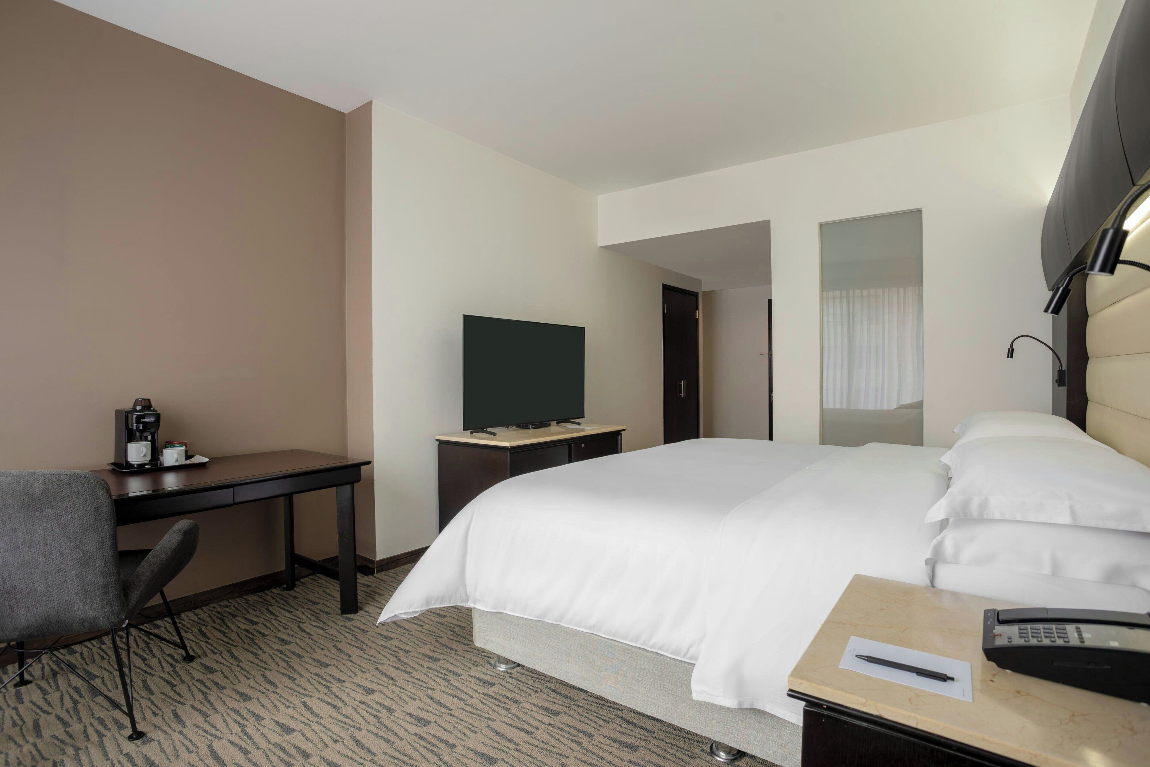 A hotel room with a bed, a desk with a chair, a television, and a bedside table with a phone and a cup.