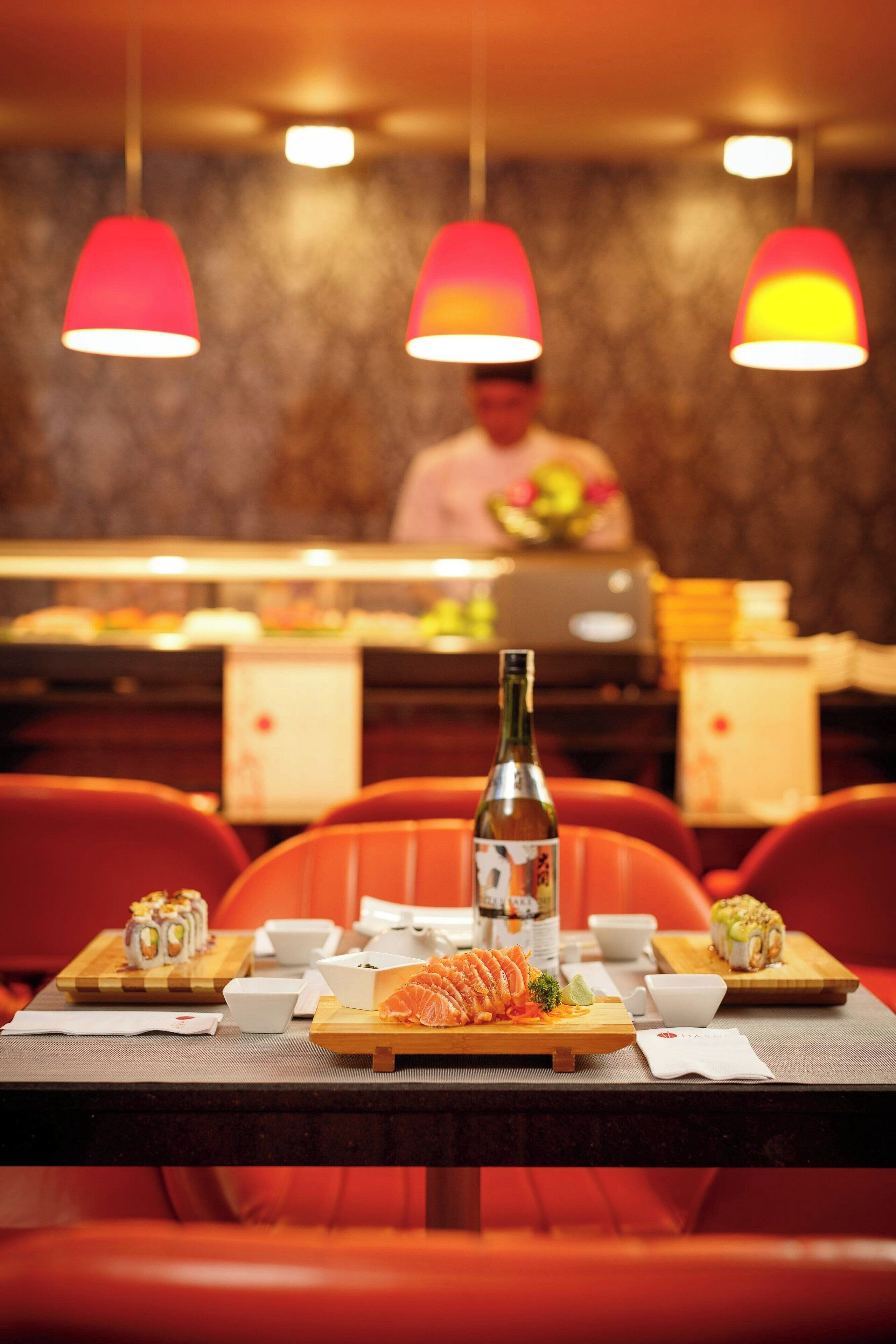 Entorn de restaurant amb un plat de sushi, una ampolla i un xef al fons.